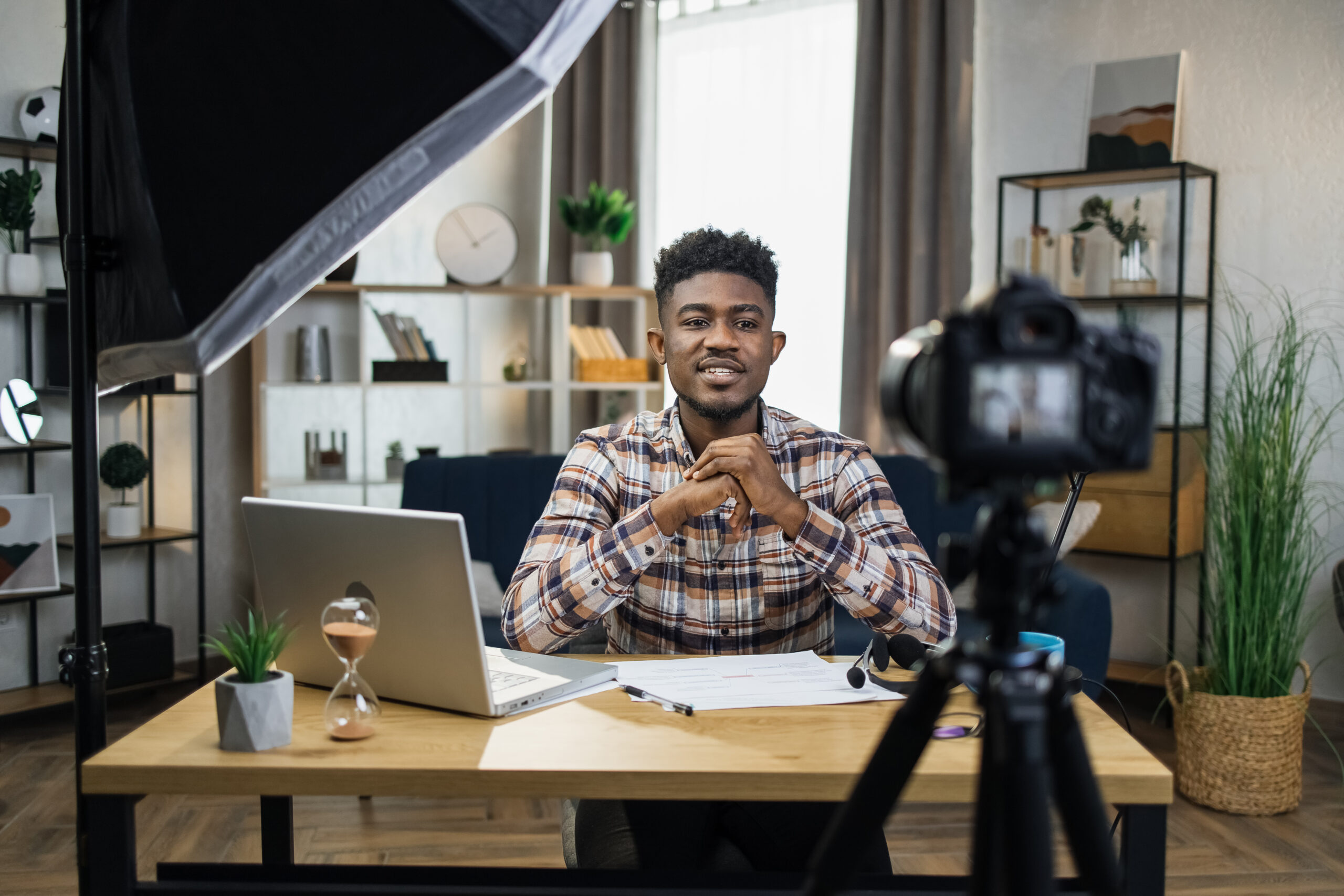 Young African man sitting at table with modern laptop and recording video on camera that fixed on tripod. Male blogger creating content for his social media.